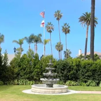 A tiered water fountain stands on a lawn surrounded by hedges and palm trees, with two flags on flagpoles visible in the background under a clear sky.