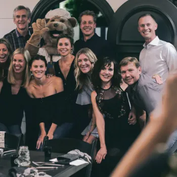 A group of adults poses together indoors, smiling for a photo; a person in a dog mascot costume stands in the back.