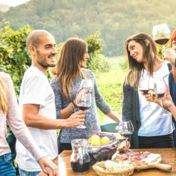 A group of people stand outdoors around a table with food and wine, talking and laughing together in a vineyard setting.