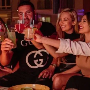 A group of seven people sitting closely together indoors, raising their drinks and smiling, with food and drinks on the table in front of them.