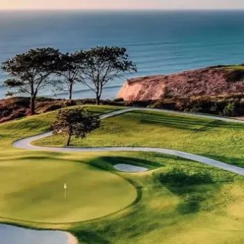 A golf course with a green, sand trap, and winding path overlooks the ocean and cliffs under a clear sky at sunset.