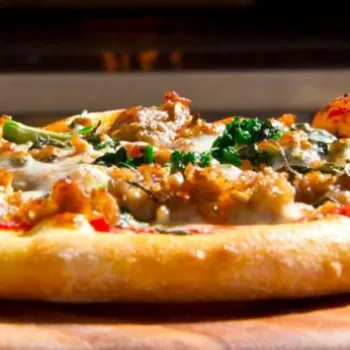Close-up of a freshly baked pizza topped with sausage, melted cheese, and green vegetables on a wooden surface.