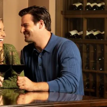 A man and woman sit at a wooden table, smiling and holding glasses of red wine, with shelves of wine bottles in the background.