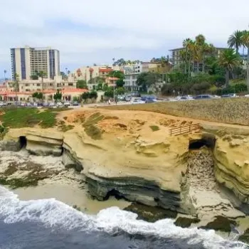 A coastal scene with sandy cliffs, people on the beach, ocean waves, and buildings, including palm trees and a tall hotel, overlooking the shoreline.