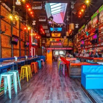 Colorful, modern bar with exposed brick walls, blue booths, multicolored stools, vibrant wall art, and overhead lighting, leading to a bar counter at the far end.