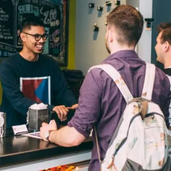 Two men with backpacks stand at a reception desk, speaking to a smiling staff member in a casual indoor setting.