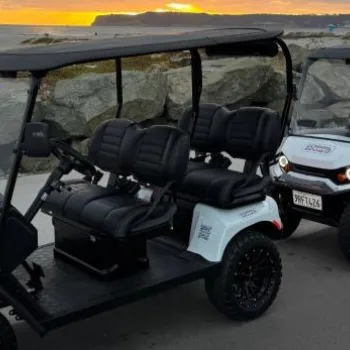 Two white golf carts with black seats are parked on a paved road by large rocks, with a beach and ocean visible at sunset in the background.
