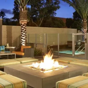Outdoor lounge area with cushioned seating around a fire pit, beside a swimming pool, in the evening with palm trees and ambient lighting.