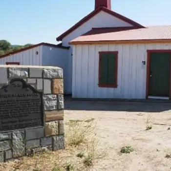 A historic white wooden building with red trim stands behind a stone marker with a plaque, set in a dry, rural landscape under a clear sky.