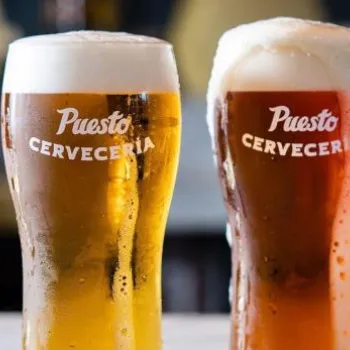 Three glasses of beer with varying colors and foam heads, each labeled "Puesto Cerveceria," are placed side by side on a table.