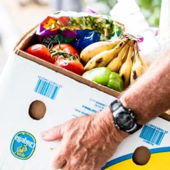A person wearing a watch carries a box filled with groceries, including bananas, tomatoes, and packaged items, on a street with cars in the background.
