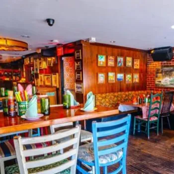 A brightly lit restaurant with colorful chairs and tables, exposed brick walls, framed artwork, and a Cuban flag displayed on the wall.