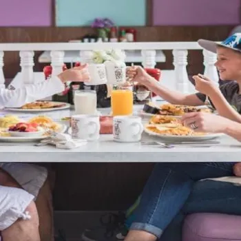 A family of four sits in a diner booth, eating breakfast together. Plates of food, a glass of orange juice, and coffee mugs are on the table.