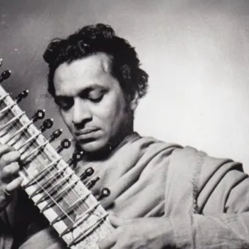 A man in traditional attire plays a sitar, focusing intently on the instrument in a black and white photograph.