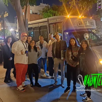 A group of nine people stand smiling in front of a Haunted San Diego Ghost Tours bus at night in an urban setting.