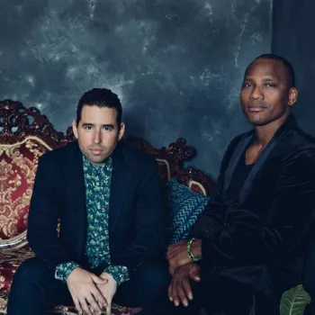 Two men sit side by side on an ornate red and gold sofa against a dark, textured wall. Both wear dark suits; one has a floral shirt, the other wears all black.