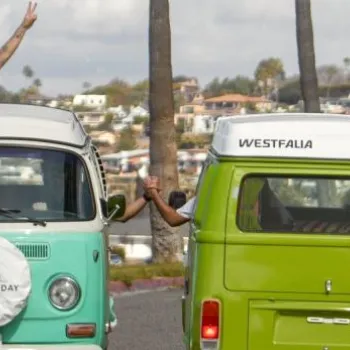 Two Volkswagen vans are parked on a street; people are leaning out of the vans, smiling and waving, with houses and palm trees visible in the background.