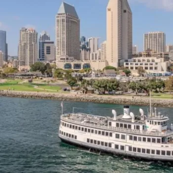 A white passenger boat sails in the foreground with the skyline of downtown San Diego and waterfront park visible in the background under a clear sky.