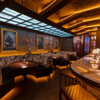 A warmly lit bar with round tables, patterned booth seating, framed paintings on the wall, and a curved counter with cushioned stools.