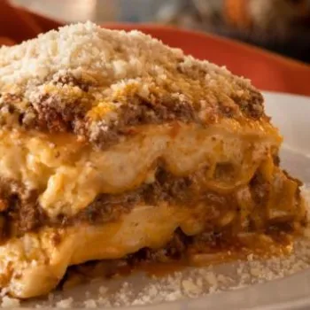 A slice of lasagna with layers of meat, cheese, and sauce is served on a white plate, topped with grated cheese.