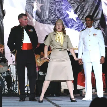 Three people in military uniforms perform on stage in front of a large American flag backdrop, with a band playing in the background.