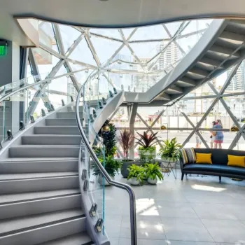 Curved staircase with glass railing inside a modern building, large geometric glass windows, potted plants, and a black couch with yellow pillows.