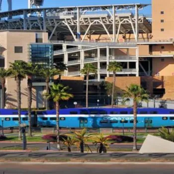 A blue and white Coaster train passes in front of a hotel and a large stadium with palm trees lining the street.