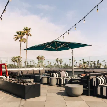 Rooftop patio with black and white striped lounge seating, a fire pit, umbrellas, string lights, and palm trees in the background under a partly cloudy sky.