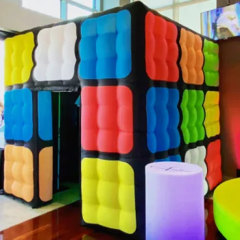 Large inflatable structure designed to look like a Rubik's Cube in an indoor event space, with people standing nearby and city buildings visible through the windows.