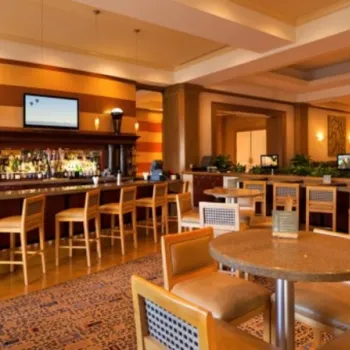 Modern hotel bar and lounge area featuring a long bar with bottles and a TV, high chairs, round tables with chairs, and contemporary decor with warm lighting.