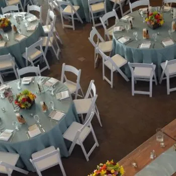 Round tables with blue tablecloths, white chairs, and colorful floral centerpieces set for an event, with a long rectangular table on the side.