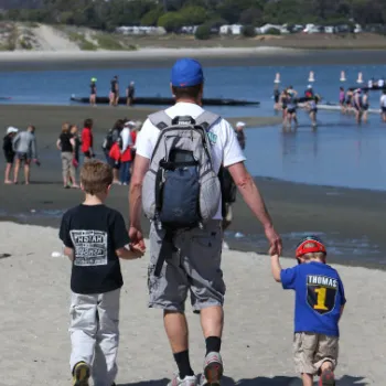 An adult and two children walk on a sandy beach toward the water, holding hands, with other people visible near the shore in the background.