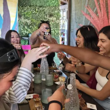 A group of women sit around a table, smiling and raising shot glasses in a toast at an outdoor restaurant or bar.
