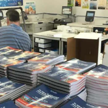A person works in a print shop surrounded by stacks of spiral-bound booklets, paper piles, and printing equipment.