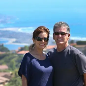 A man and woman stand together outdoors smiling, with a coastal town, hills, and ocean visible in the background.