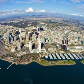 Aerial view of a coastal city with a dense downtown area, marinas, piers, and surrounding suburbs under a blue sky with scattered clouds.