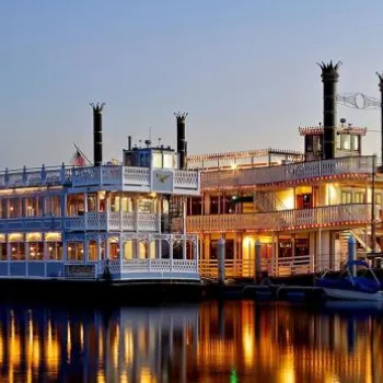 A brightly lit paddlewheel riverboat is docked on calm water at dusk, with sailboats moored nearby and palm trees in the background.