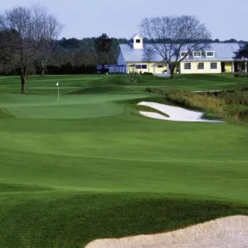 A golf course with sand bunkers, a putting green, and a clubhouse in the background surrounded by trees and a pond.