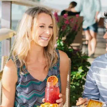 A woman and a man sit at an outdoor table by the water, smiling and enjoying food and drinks on a sunny day.