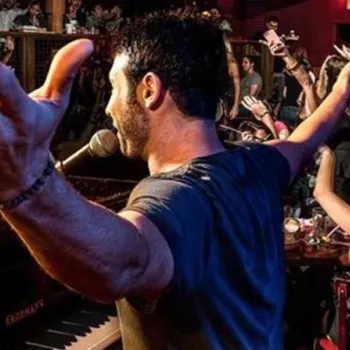 A man stands at a piano with arms outstretched, singing into a microphone, as a lively audience cheers and raises their hands in a crowded, dimly lit venue.