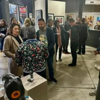 A group of people socialize and view artwork at a gallery exhibition, with paintings displayed on the walls and sculptures on pedestals.