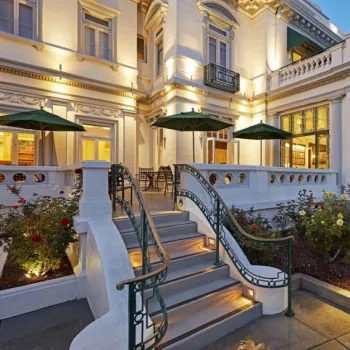 A large, elegant building with white exterior walls, ornate railings, steps leading to a terrace, green umbrellas, and landscaped garden beds at dusk.