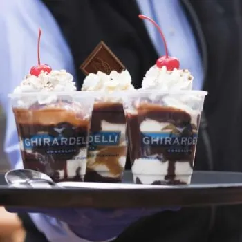 A server holds a tray with three Ghirardelli sundaes topped with whipped cream, chocolate pieces, and cherries, along with a spoon, at an outdoor seating area.