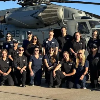 A group of people in matching black shirts pose in front of a military helicopter on a sunny day at an airfield.