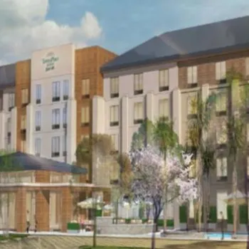 A four-story hotel building with a sign reading "Homewood Suites," surrounded by trees and landscaping under a partly cloudy sky.