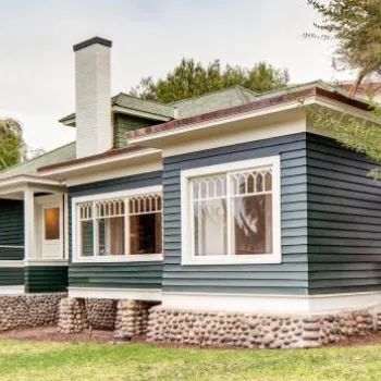 A single-story house with dark green siding, white trim, large windows, and a stone foundation, surrounded by trees and grass.