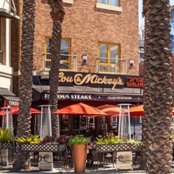 Outdoor restaurant seating with red umbrellas and palm trees, located near the entrance to San Diego’s Gaslamp Quarter, with historic and modern buildings in the background.