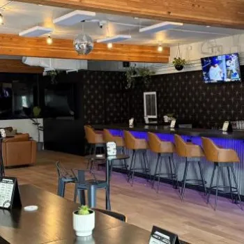 Modern bar with wooden beams, blue-lit counter, high chairs, several TV screens, and a seating area with tables and couches.