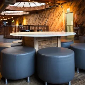 A modern lounge area with round tables, gray upholstered stools, wood-paneled walls, and string lights. The space is empty with a warm, ambient atmosphere.