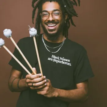 Smiling person with dreadlocks holds three mallets and wears a black t-shirt that says "Black Women are Superheroes" in white text, standing against a brown background.
